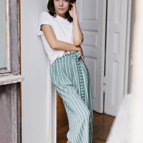 Barefooted young lady in cozy clothes posing at home. Brunette elegant girl in white t-shirt chilling in weekend in her apartment.
