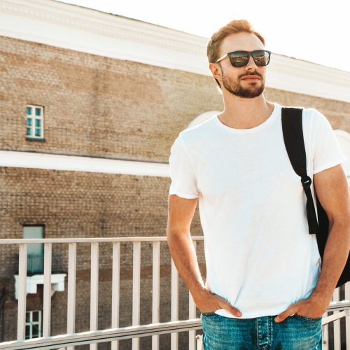 Handsome smiling  hipster lambersexual model.Stylish man dressed in white T-shirt and jeans. Fashion male posing on the street background near fence at the beach with schoolbag in sunglasses