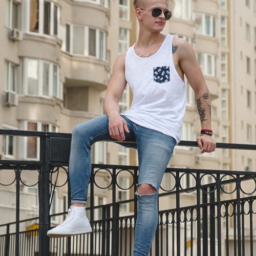 Stylish young man with tattoos and trendy hairdo, wearing sunglasses, white shirt and jeans relaxing while sitting in the city on buildings background. Male watching city life.