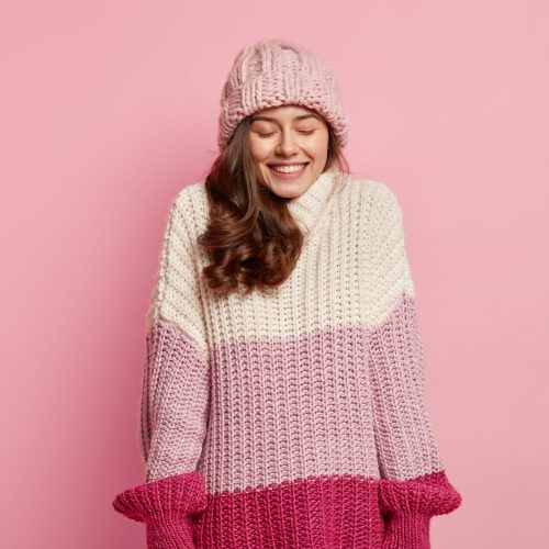 Isolated shot of pleased woman feels enjoyment from something positive, wears knitted hat and sweater, smiles gently, models against pink background, satisfied with good news. Winter time concept